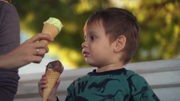 Η μαμά και ο γιος τρώνε παγωτό. Μαμά και παιδί. Το παιδί τρώει παγωτό. Ένα μικρό αγόρι με τη μητέρα του στον πάγκο τρώει παγωτό, αλλά το παιδί θέλει να ανταλλάξει παγωτό με τη μητέρα του. — Αρχείο Βίντεο