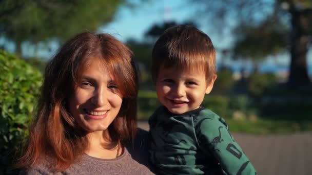 Close-up van een twee jaar oud zoontje in zijn armen en een jonge moeder. De jongen zwaait met zijn hand naar de camera. Een jonge moeder met een klein kind in haar armen kijkt naar de camera en glimlacht. — Stockvideo