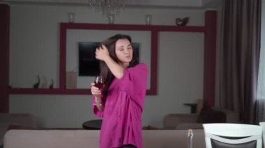 Genç bir kız ev hanımı zor bir günün ardından evde dinleniyor, müzik dinliyor, kırmızı şarap içiyor ve dans ediyor. Evde dans eden ve bardaktan şarap içen genç bir kızın portresi. 4k