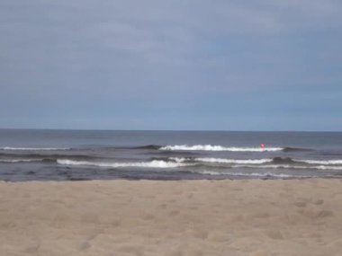 Kum plajında köpüklü Baltık deniz dalgaları, suda ve uzakta kırmızı güvenlik şamandırası, beyaz bulutlu mavi gökyüzü