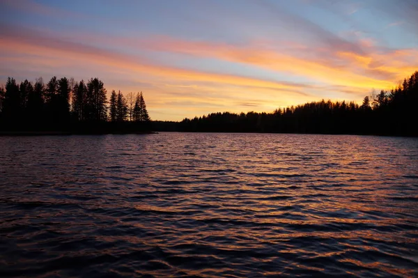 Sunset on the lake. On the horizon a dark forest. The sun had gone down behind the treetops. The sky is painted blue, yellow and orange. The water is dark blue with orange reflections.