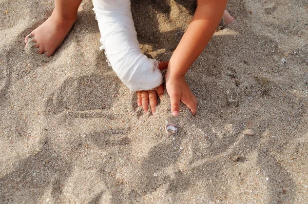 Kind mit Gips an der Hand spielt im Sand am Strand am Meer. — Stockfoto