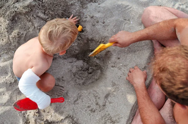 Papa und Baby mit Gips an der Hand spielen im Sand am Strand am Meer. Gesundheit und Erholung — Stockfoto