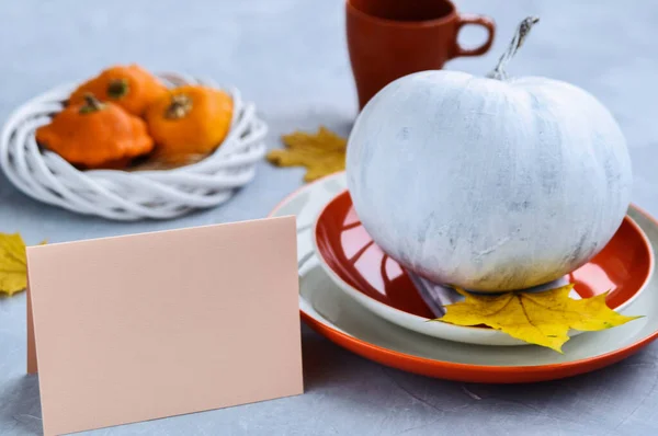 Halloween or Thanksgiving card on the table decorated with Pumpkin painted in white, orange plate and yellow leaves.