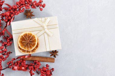 İçinde Holly meyveleri, kırmızı kurdele, kurutulmuş portakal dilimleri, tarçın ve gri beton arka planda beyaz hediye kutusu olan Noel kartı