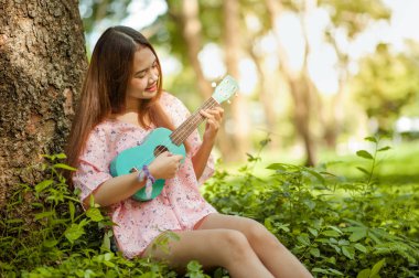 şapka lı asyalı bayan yaz aylarında ukulele bossanova müzik çalar. Genç sevimli kadın açık havada müzik çalıyor. Mutlu bir genç kız bir ağacın altında ukulele oynamaktan hoşlanıyor. Arka planda güzel doğa.