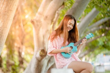 Yaz aylarında şapka oyun ukulele Bossanova müzik ile Asya Bayan. Genç Sevimli kadın açık havada müzik oynuyor. Mutlu bir genç kız bir ağacın altında ukulele oynarken hoşlanır. 