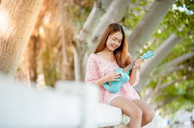 Yaz aylarında şapka oyun ukulele Bossanova müzik ile Asya Bayan. Genç Sevimli kadın açık havada müzik oynuyor. Mutlu bir genç kız bir ağacın altında ukulele oynarken hoşlanır. 