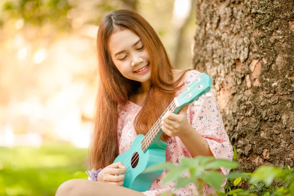 帽子をかぶったアジアの女性は 夏の時間にウクレレボサノバ音楽を演奏します 屋外で音楽を演奏する若いかわいい女性 幸せな若い女の子は木の下でウクレレを楽しんでいます 背景の美しい自然 — ストック写真
