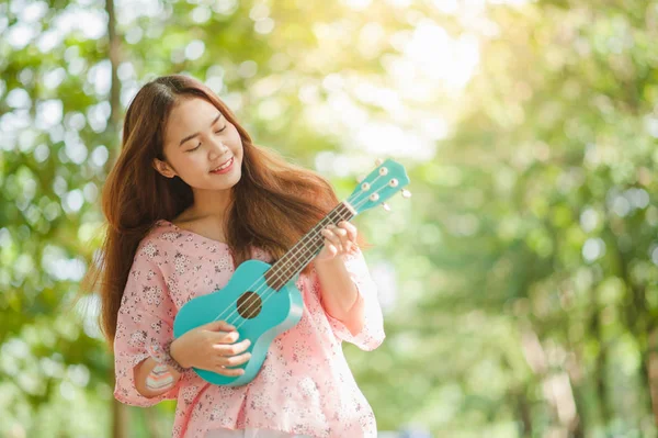 asian lady with hat play ukulele bossanova music in summer time.Young cute woman playing music outdoors.A happy young girl enjoys playing ukulele under a tree. Beautiful nature in the background.