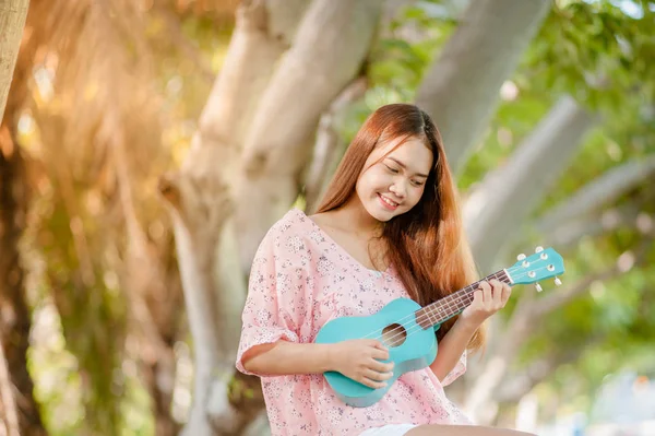 asian lady with hat play ukulele bossanova music in summer time.Young cute woman playing music outdoors.A happy young girl enjoys playing ukulele under a tree.