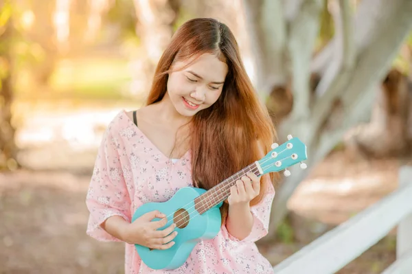 asian lady with hat play ukulele bossanova music in summer time.Young cute woman playing music outdoors.A happy young girl enjoys playing ukulele under a tree.