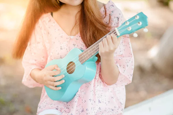 asian lady with hat play ukulele bossanova music in summer time.Young cute woman playing music outdoors.A happy young girl enjoys playing ukulele under a tree.