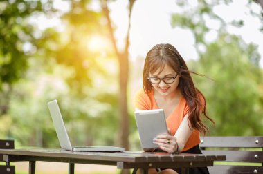 Genç ve güzel Asyalı bir kadın laptopla / internetten / açık hava parkında alışveriş yapıyor, sabahları gülümsüyor / ferahlatıyor ve rahatlatıyor, serbest çalışan iş kadını yaşam tarzı kavramı.