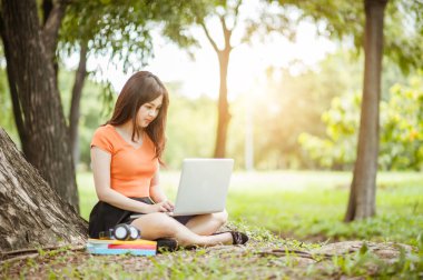 Genç ve güzel Asyalı bir kadın laptopla / internetten / açık hava parkında alışveriş yapıyor, sabahları gülümsüyor / ferahlatıyor ve rahatlatıyor, serbest çalışan iş kadını yaşam tarzı kavramı.