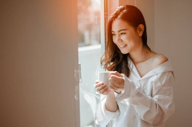 Elinde bir fincan kahve tutan bir kadın. Penceresinde güneş doğarken evde kahve içen bir kadın. .