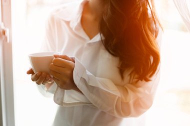 Elinde bir fincan kahve tutan bir kadın. Penceresinde güneş doğarken evde kahve içen bir kadın. .