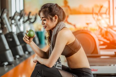 Spor salonunda antrenman yaptıktan sonra dinlenen atletik bir kadın. Koşu bandında oturup elma yiyor.. 