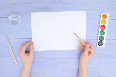 Kağıt ve suluboya boya menekşe ahşap arka plan üzerinde beyaz bir boş levha ile eller fırça ile kızın. Yukarıdan görüntülemek