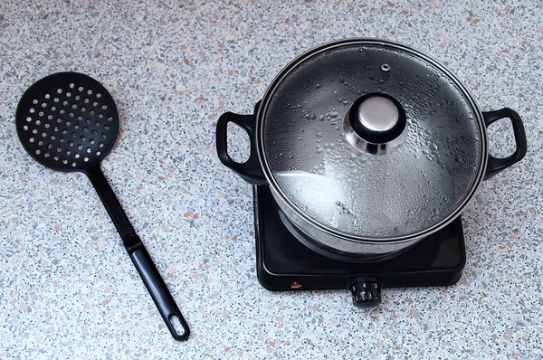 Saucepan on a small electric stove with a lid for a couple. On the table is a plastic black skimmer spoon. View from above.