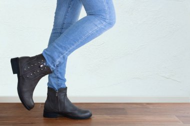 Mavi kot pantolonlu ve kahverengi çizmeli bir kızın bacakları. Beyaz bir duvarın arka planına karşı.