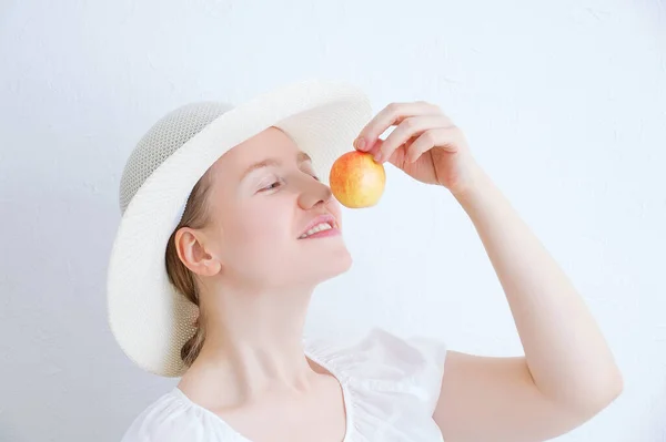 Caucasian young woman in white tunic and straw hat smiling with an apple in her hands against a white wall.