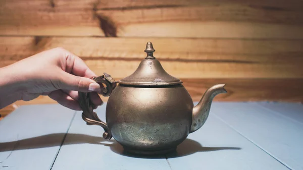 Woman hand holding handle of vintage copper teapot kettle with blemishes and marks, sitting in blue wooden table.