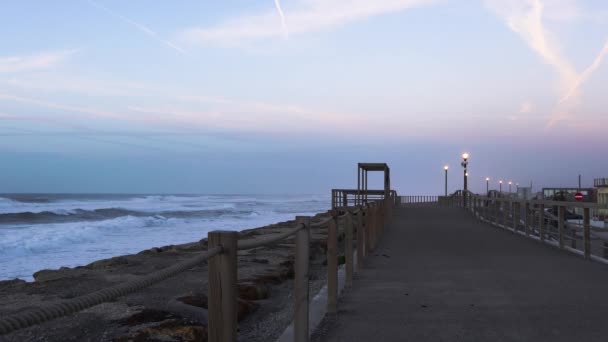 Havsvågor kraschar nära stranden, bredvid tomma gångväg upplyst av gatubelysning. — Stockvideo