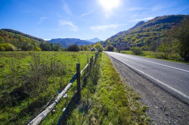 Brasov yakınlarındaki Karpatya dağlarında bulutlu gökyüzü ve güneş ışığıyla çayırlar ve dağlar arasında uzanan boş asfalt köy yolunun ilkbahar manzarası
