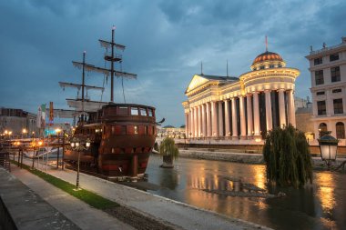 Skopje, Republic of Macedonia - 16 October, 2019: The archaeological Museum of Macedonia and the Bridge of Civilizations at night. Old ship in the river. Beautiful light clipart