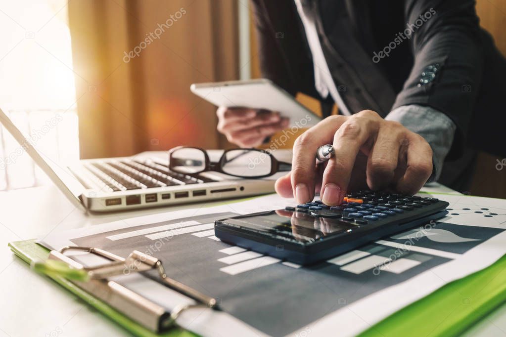 businessman hand working with finances about cost and calculator and laptop with tablet on withe desk at office in morning light