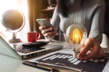  Ofiste dizüstü bilgisayar ve para destesi kullanan bir iş adamı ampulü tutuyor. Sabah ışığında enerji ve muhasebe finans kavramının korunması fikri