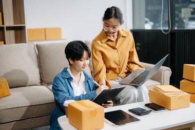 Başlangıçta küçük işletme KOBİ 'si, girişimci sahibi dizüstü bilgisayar ve tablet kullanan iki kadın paket ürün kutusu hazırlamak için online alışveriş siparişi alıyor ve kontrol ediyor.