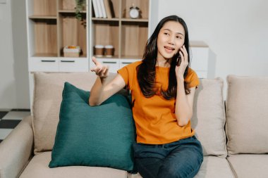 Genç Asyalı kadın evdeki kanepede dinleniyor ve akıllı telefonla konuşuyor.