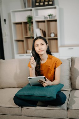 Genç Asyalı kadın evdeki kanepede dinleniyor ve dijital tablet kullanıyor.