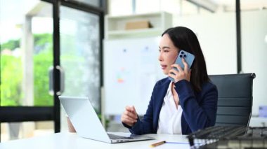 Bilgisayarda çalışırken akıllı telefondan konuşan genç bir iş kadını.