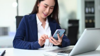 Genç iş kadını bilgisayarla masada otururken akıllı telefon kullanıyor.