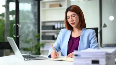 Genç iş kadını modern ofis masasında otururken notlar yazıyor.