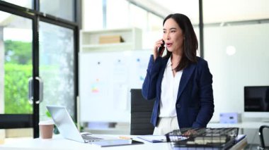 Genç iş kadını modern ofiste dururken telefonla konuşuyor.