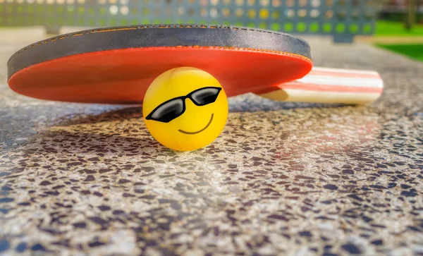 Tennis table paddle with a emoticon ball below it on a stone tennis table in a park. Loving sport concept.