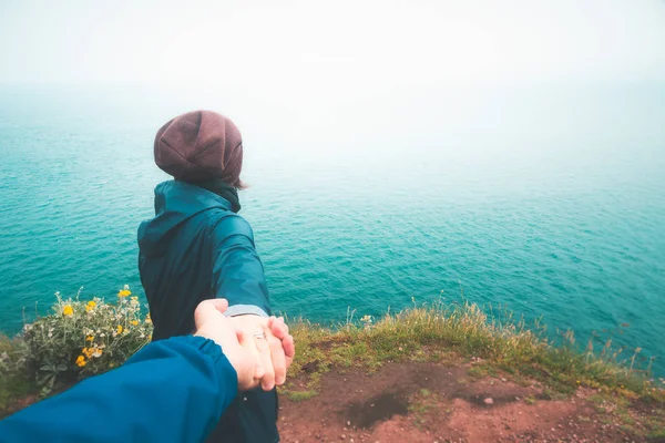 Follow me to the sea. Girl taking hand of her boyfriend leading him to the cliff edge overlooking the sea on a moody and rainy day. Outdoors, nature, travel, freedom, hiking concepts.