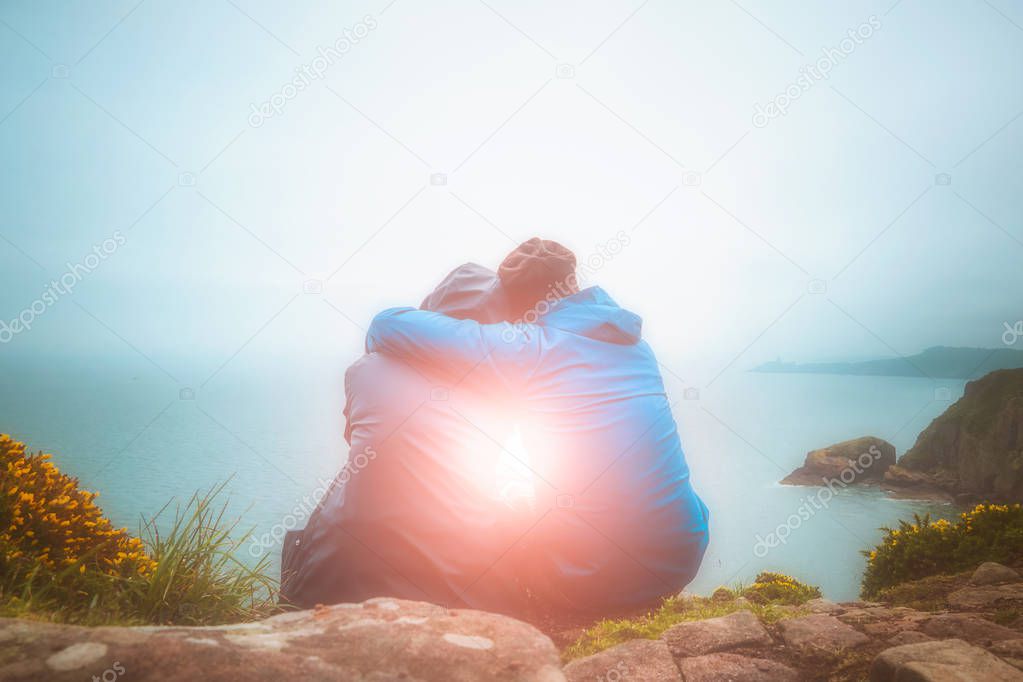 Rear or back view of young lovers sitting against each other and hugging on a cliff edge looking at a scenic seascape with light burst. Love, romance, outdoors, nature, freedom concepts.