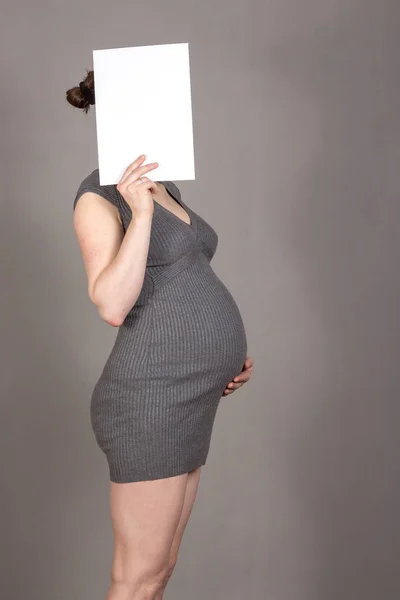 Schwangere auf neutralem grauen Hintergrund mit einem Stück Papier. Platz für die Inschrift. — Stockfoto