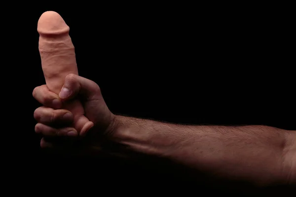 Adult present. Artificial penis in mans hand on dark background.