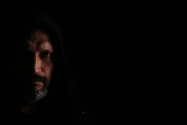 Kapüşonlu gri sakallı, siyah arka planda keskin gölgeleri olan acımasız bir adam. Kopyalama alanı.