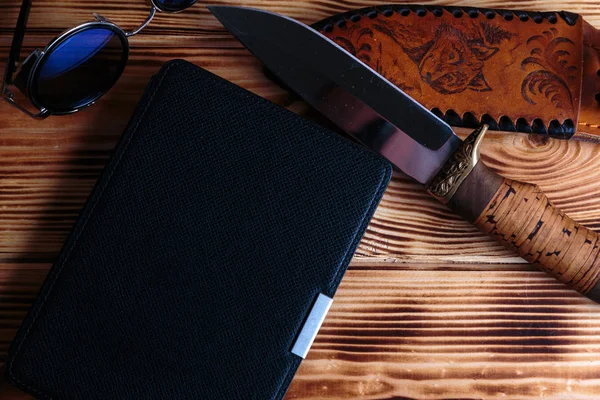 Electronic book  in case with hunting knife and sunglass on wooden background. Adventure or detective book concept.