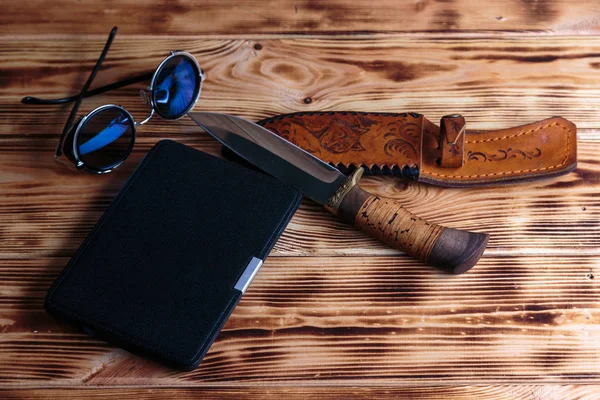 Electronic book  in case with hunting knife and sunglass on wooden background. Adventure or detective book concept.