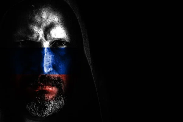 A brutal man with a gray beard, with the flag of Russia on his face, in a hood with sharp shadows on a black background. Copy space