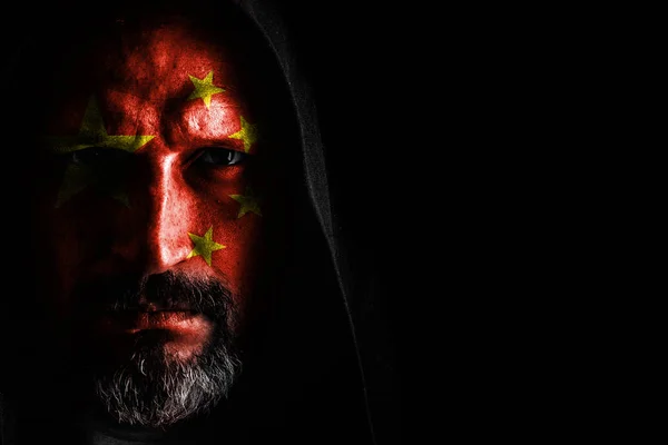 A brutal man with a gray beard, with the flag of China on his face, in a hood with sharp shadows on a black background. Copy space — Stock Photo, Image
