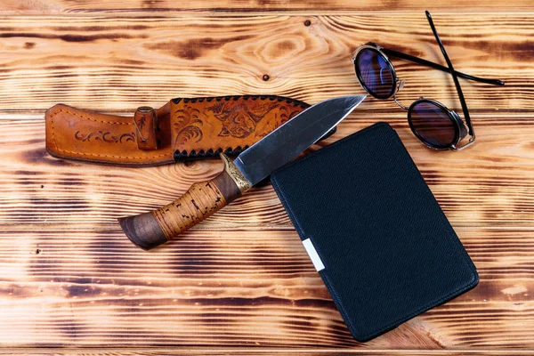 Electronic book  in case with hunting knife and sunglass on wooden background. Adventure or detective book concept.